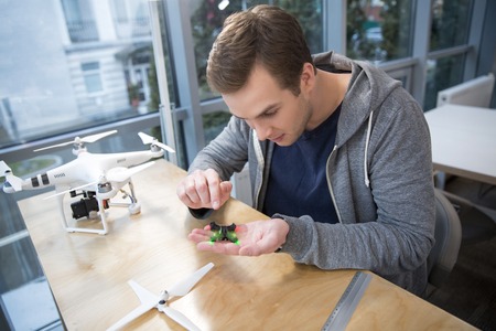 Portrait of skillful young freelancer designing a small drone. He is holding the technology in his...