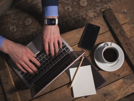 Close up of male hands typing on the laptop. The man is sitting near a table with a cup of coffee and mobile phone on itの写真素材
