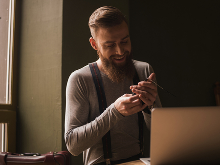 Waist up portrait of happy bearded man in retro styled clothing. He is wiping his eyeglasses and smiling. The man is sitting near the laptopの写真素材
