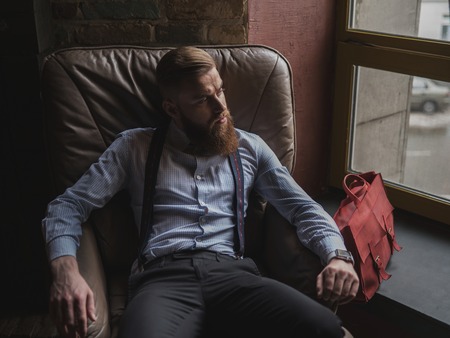 Portrait of attractive man with beard resting in armchair. He is looking trough the window pensively. The guy is wearing retro clothingの写真素材