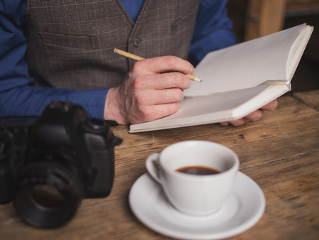 Close up of male hands making notes into the writing-pad. The man is sitting at the table near the camera. He is drinking coffeeの写真素材