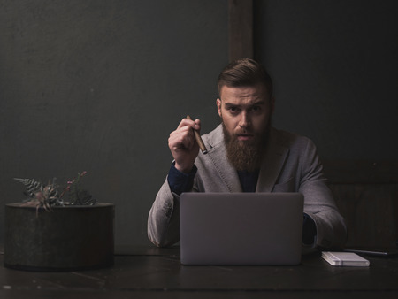 Cheerful young businessman with beard is using a laptop. He is sitting at the desk and smoking an expensive cigar. The guy is looking at camera with confidenceの写真素材