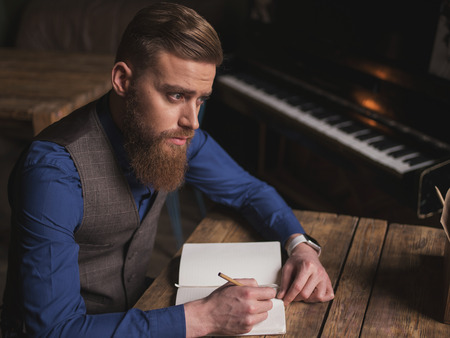 Attractive bearded poet is noting his ideas in the copybook. He is sitting at the table. The man is looking forward with aspirationの写真素材