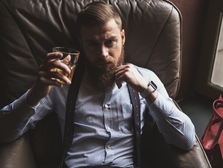 Handsome young bearded businessman is drinking expensive whisky. He is sitting in armchair and holding stretching a glass to the camera. The guy is looking forward seriouslyの写真素材