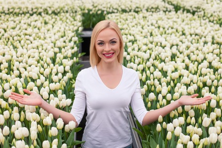 Beautiful young woman is standing in flower garden and smiling. She is posing and stretching arms on tulips happilyの写真素材