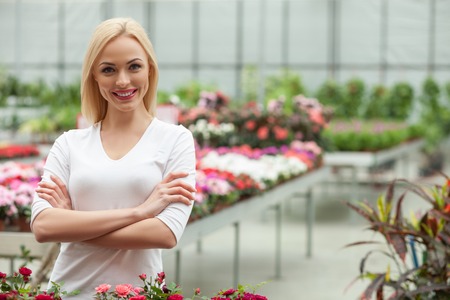 Beautiful female florist is caring of the flowers in plant nursery. She is standing and looking at camera happily. The woman is smiling. She crossed her arms with confidenceの写真素材