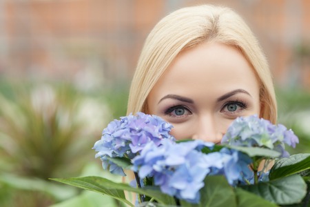 Pretty female florist is holding a flowerpot in shop. She is hiding behind the blue flowers and looking at camera with joy. Copy space in left sideの写真素材