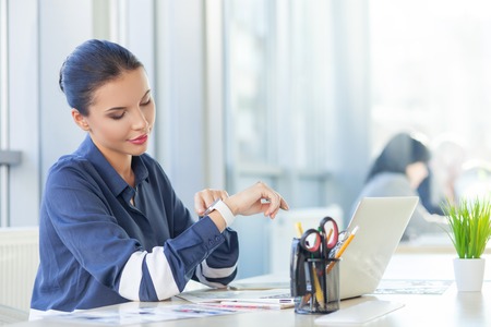 Cheerful young woman is sitting at the desk in her office. She is looking at her smart watch and smilingの写真素材