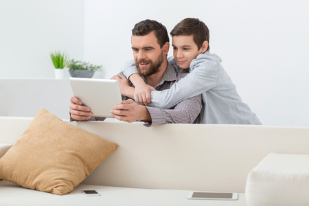 Cute father is playing game on the tablet. His son is looking at the screen and embracing him. They are smilingの写真素材