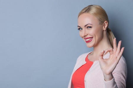 Waist up portrait of attractive blond girl showing ok sign. She is standing and smiling. Copy space in left sideの写真素材