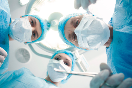 Low angle of experienced mature surgeon is operating with concentration in surgery. His colleagues are standing and watching the operation with seriousnessの写真素材