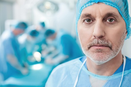 Experienced mature surgeon is looking forward seriously. He is standing in blue uniform. His...