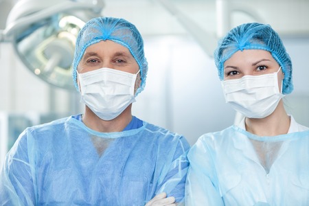 Everything is okay. Portrait of cheerful two surgeons expressing positive emotions. They are standing and looking forward with confidence. The man and woman are wearing masksの写真素材