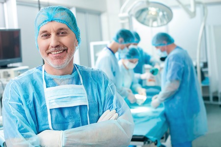 Everything will be okay. Cheerful mature surgeon is smiling and looking forward with joy. His surgical team is doing the operation on the backgroundの写真素材