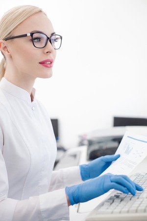 Cheerful female laboratory technician is using a computer for work. She is typing on the keyboard and smiling. The woman is holding a paper of researchの写真素材