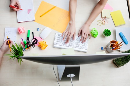 Top view close up of arms of creative team.  Female arms are typing on a computer in office. Her colleagues are drawing diagrams on papersの写真素材