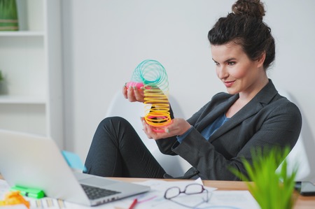 Attractive young woman calms down playing with a toy spiral. She is sitting in her office and relaxing. The woman is smilingの写真素材