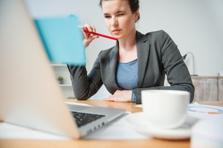 Low angle of beautiful young businesswoman is working in office. She is looking at the laptop and screwing up her eye pensively. The lady is sitting at the desk near a cupの写真素材