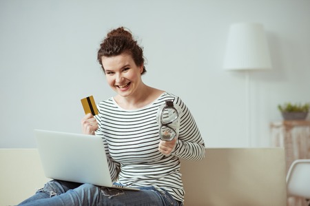 Cheerful girl is holding a diving mask and credit card. She is sitting on sofa and laughing. The lady is carrying a laptop on her kneesの写真素材