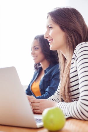 Pretty two young women are listening to someone with interest. They are sitting at the desk near a...