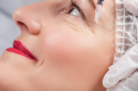 Close up of face of senior woman getting botox injection in eye area. She is smiling gently. The needle of syringe is touching female wrinkleの写真素材