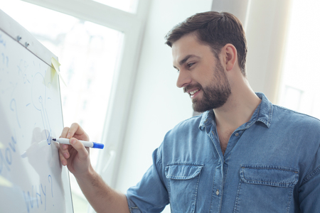 Attractive young businessman is writing his ideas on the board. He is standing and smiling in officeの写真素材