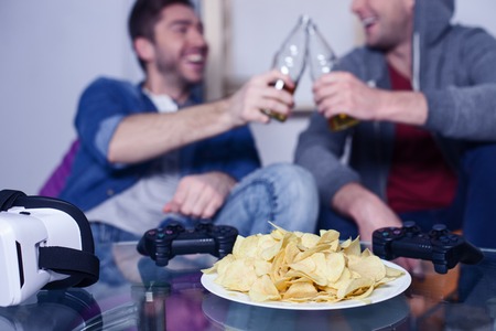 Handsome young men are drinking beer and talking. They are clicking bottles and smiling. Focus on chips and play-station on the tableの写真素材