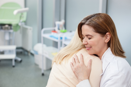 Pretty female gynecologist is congratulating woman with her pregnancy. She is embracing her and smiling. They women are sitting in hospital officeの写真素材