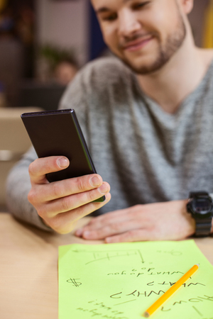 Cheerful man is holding a mobile phone and messaging. He is sitting at desk near a sheet of paper and pencil. The student is smilingの写真素材