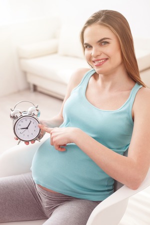 Cheerful young expectant mother is pointing finger at clock. She is sitting and smiling
