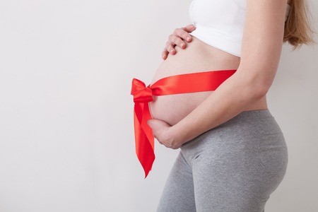 Midsection of pregnant woman standing in profile. Her abdomen is wrapped with red ribbon and bow. The lady is embracing her belly gently. Isolated and copy space in left sideの写真素材