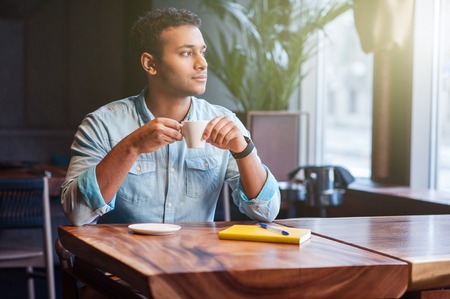 Handsome young man is enjoying coffee in cafe. He is looking through window pensively and dreamingの写真素材