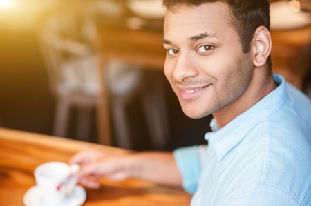 I like resting here. Waist up portrait of cheerful young man looking at camera and smiling. He is sitting at table and drinking coffee in cafeの写真素材