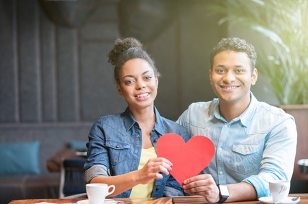 We love each other. Attractive young man and woman are sitting at table in cafe. They are embracing and smiling. The lovers are holding card in shape of heartの写真素材