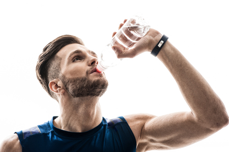 Waist up portrait of thirsty handsome young sportsman drinking water from bottle. He is standing in sportswear. Isolated on backgroundの写真素材