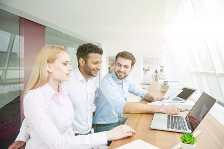 Friendly team is working together with joy. They are looking at laptop and smiling. Men and woman...