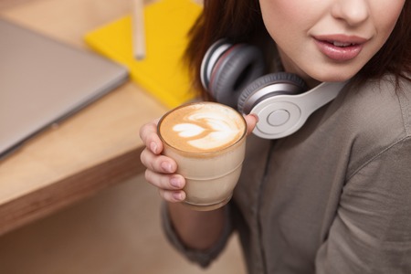Cheerful girl is making break after studying. She is holding a cup of latte and smiling. The lady is sitting near a desk with laptopの写真素材