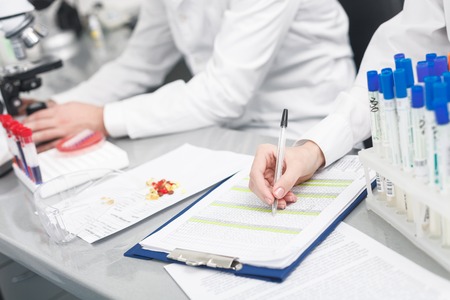 Close up of hands of young researchers working with samples. Woman is sitting at desk and making notes. Man is looking into microscopeの写真素材