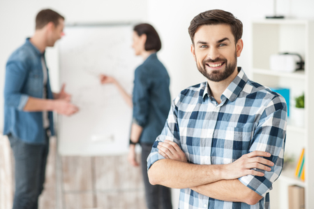Waist up portrait of attractive young man looking at camera with confidence and smiling. His colleagues are standing and talking on backgroundの写真素材