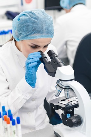 Cheerful female researcher is analyzing substance in lab. She is looking at microscope with...