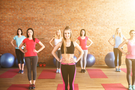 Attractive young women are doing exercise in gym. They are standing with arms akimbo. The ladies are smilingの写真素材