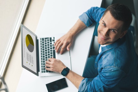 Top view of attractive young man using a laptop for work. He is sitting at desk and looking up with joy. The man is smilingの写真素材