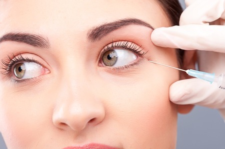 Close up of female face getting botox injection. Young woman is looking aside with concentration. Hand of doctor is holding a syringe near her eyeの写真素材