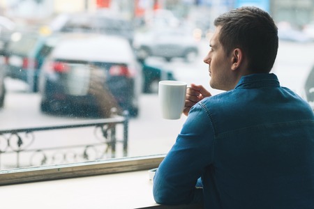 Attractive young man is drinking coffee with pleasure. He is looking at city through the window. The man is resting and dreamingの写真素材