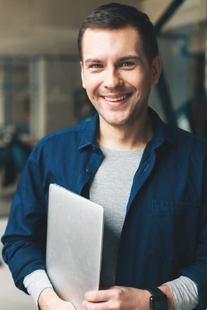 Cheerful guy is holding a laptop. He is standing and smiling