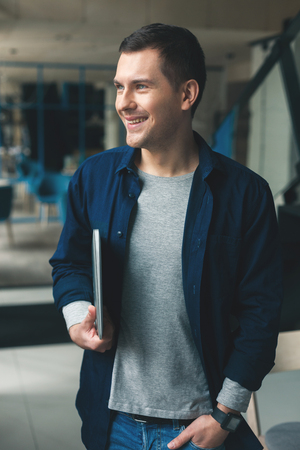 Portrait of handsome young man carrying a laptop. He is standing and putting arm in his pocket. The man is looking aside and smilingの写真素材