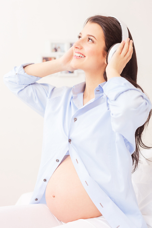 Beautiful young pregnant woman is enjoying a melody at home. She is touching headphones and smiling. The lady is sitting on bedの写真素材