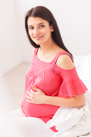Beautiful young expectant mother is touching her belly with love. She is sitting on bed and smilingの写真素材