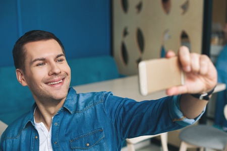 Attractive young man is making selfie in cafe. He is holding a mobile phone and smilingの写真素材