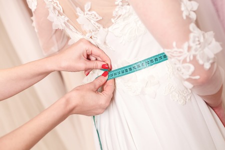 Close up of arms of tailor measuring female waist with tape-measure. Young bride is trying on a wedding dressの写真素材